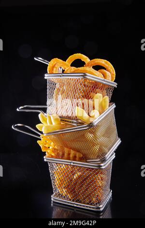 Tas de délicieuses rondelles d'oignon avec des frites et des chips servies dans de petits paniers en maille métallique sur fond noir Banque D'Images