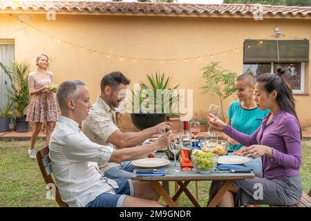 Multiculturel heureux amis riant avec un beau sourire autour de la table de déjeuner. Tandis que la femme apporte la nourriture à la maison patio. Banque D'Images