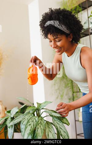 Jeune femme biraciale souriante avec cheveux afro versant de l'eau sur les plantes tout en étant debout à la maison Banque D'Images