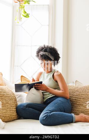 Jeune femme biraciale souriante avec cheveux afro écrivant dans un journal tout en étant assise sur un canapé près de la fenêtre Banque D'Images