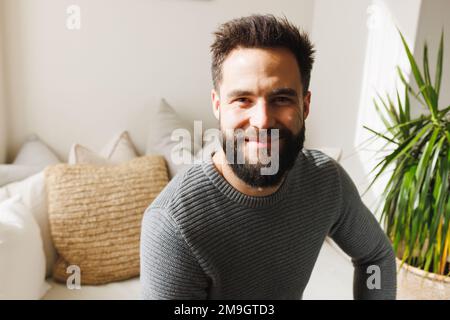 Portrait d'un jeune homme biracial à barbe souriante assis sur un canapé dans le salon au-dessus du mur, espace de copie Banque D'Images