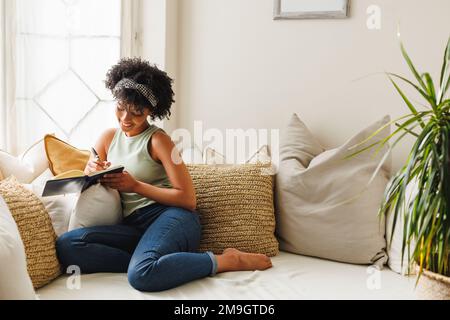 Jeune femme biraciale souriante avec cheveux afro écrivant dans un journal tout en étant assise par la fenêtre sur le canapé Banque D'Images