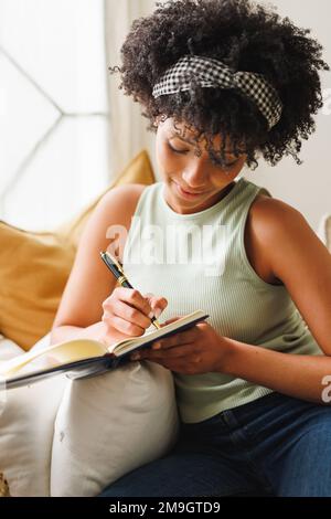 Jeune femme biraciale souriante avec cheveux afro écrivant dans un journal tout en étant assise sur un canapé Banque D'Images