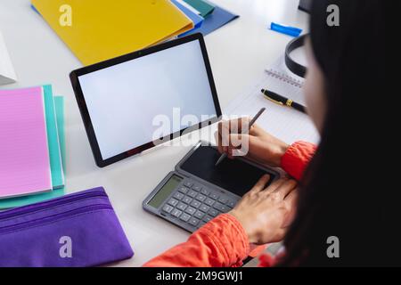 Image d'une femme biraciale utilisant une tablette avec espace de copie à l'écran et une calculatrice assise au bureau Banque D'Images