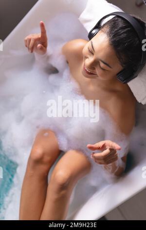 Image d'une femme biraciale se détendant avec un casque écoutant de la musique dans une baignoire dans un bain moussant Banque D'Images