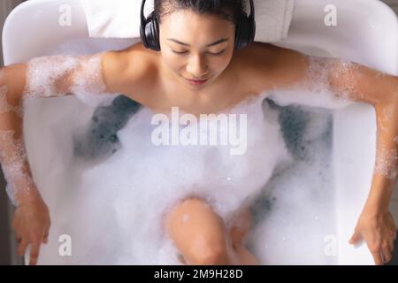 Image d'une femme biraciale se détendant avec un casque écoutant de la musique dans une baignoire dans un bain moussant Banque D'Images