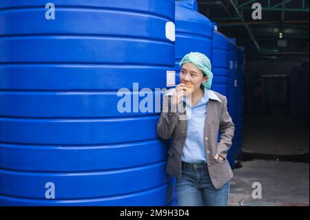 Jeune femme superviseur asiatique debout mangeant du pain avec la hâte pendant la pause déjeuner dans l'entrepôt de réservoir d'eau dans l'usine de transformation Banque D'Images