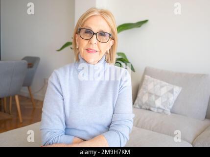 Portrait d'une belle femme âgée souriante avec des cheveux blonds assis sur le canapé à la maison. Banque D'Images