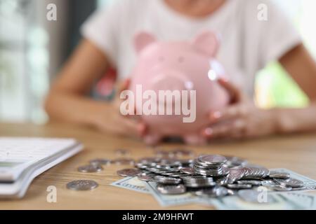 Billets et pièces de monnaie sur table et femme floue avec la banque de porc en arrière-plan. Banque D'Images