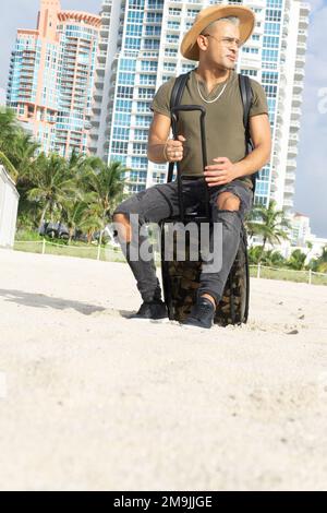 Gros plan d'un jeune homme faisant une pause sur son voyage. Un homme assis sur sa valise de transport sur la plage. South Beach, Miami. Concept voyage et été Banque D'Images