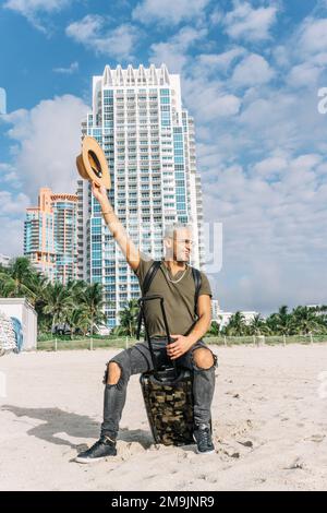 un jeune latino avec un look moderne et un chapeau sur la plage de miami va en vacances et se déplace dans la valise comme un cow-boy. Concept de vacances Banque D'Images