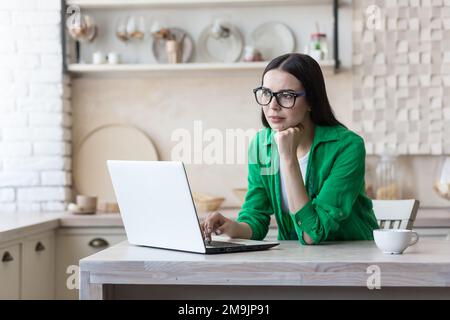 Une jeune femme contrariée assise à la maison dans la cuisine, devant un ordinateur portable. Tapez sur le clavier, recherchez, vérifiez. Elle a bien posé la tête sur sa main. Banque D'Images