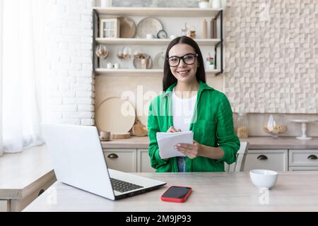 Portrait d'une jeune femme enseignante en lunettes assise à la maison dans la cuisine avec ordinateur portable. Fonctionne à domicile en ligne. Il tient un ordinateur portable avec un stylo dans ses mains, souriant à l'appareil photo. Banque D'Images