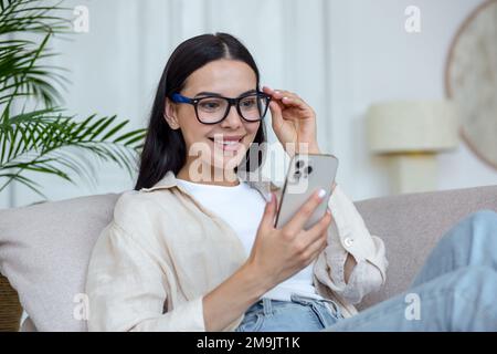 Une jeune femme heureuse en quarantaine se trouve sur le canapé à la maison et communique avec des amis, des parents, des parents, des parents par le biais d'un appel vidéo à partir du téléphone. Il sourit, dit bonjour, tient ses lunettes dans la main. Banque D'Images