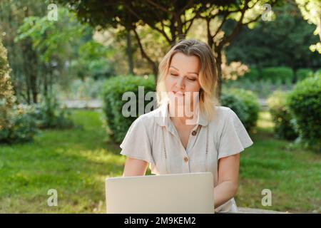 une belle femme mûre en lunettes travaille sur un ordinateur à une table blanche dans la nature et passe sa journée productive Banque D'Images