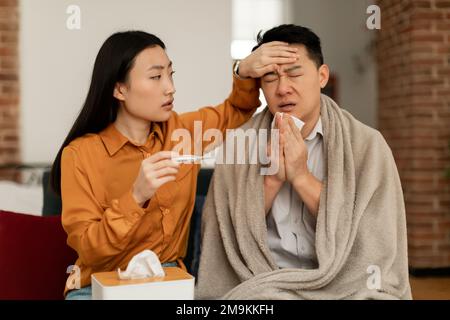Femme asiatique traitant mari malade, tenant thermomètre et touchant le front de l'homme, assis sur un canapé à la maison Banque D'Images