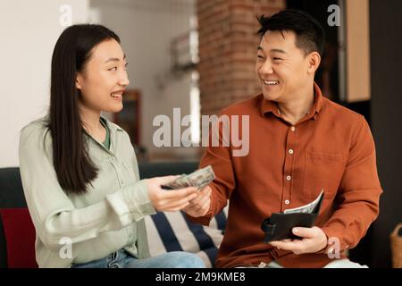 Un riche mari mature de corée donne de l'argent à sa jeune femme, assis ensemble sur un canapé à la maison Banque D'Images