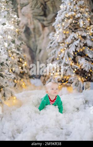 Un joli petit garçon rampant sur le sol, au milieu de la neige artificielle et des arbres de Noël décorés de guirlandes. Banque D'Images
