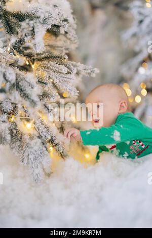 Un joli petit garçon marche sur le sol au milieu de la neige artificielle et touche les guirlandes lumineuses sur l'arbre de Noël. Banque D'Images