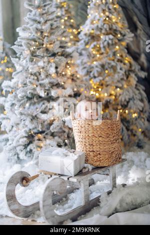 Le joli petit garçon est assis à l'intérieur d'un panier en osier sur un traîneau, au milieu de la neige artificielle et des arbres de Noël décorés de guirlandes lumineuses. Il y a Bo cadeau de Noël Banque D'Images