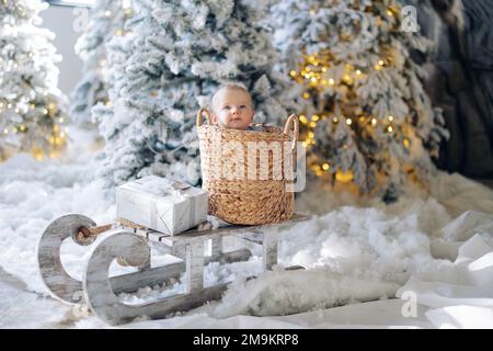 Le joli petit garçon est assis à l'intérieur d'un panier en osier sur un traîneau, au milieu de la neige artificielle et des arbres de Noël décorés de guirlandes lumineuses. Il y a Bo cadeau de Noël Banque D'Images