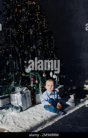 Un joli petit garçon joue sur le sol près des boîtes-cadeaux de Noël et de l'arbre de Noël avec des décorations et des guirlandes lumineuses. Banque D'Images