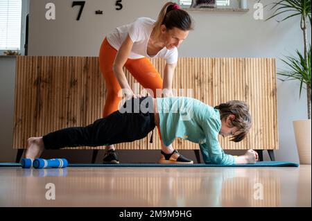 Jeune femme, une mère, aidant son fils à exécuter une position correcte de planche tout en faisant de l'exercice ou de l'exercice à la maison dans un salon sur un tapis. Banque D'Images