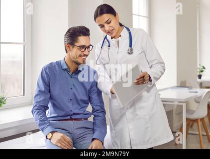 Une femme médecin professionnelle sourit et montre des résultats d'examen positifs à un homme jeune et joyeux. Banque D'Images