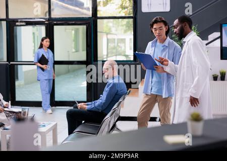 Un médecin afro-américain explique l'expertise de la maladie à un patient asiatique pendant la consultation de visite de contrôle dans la zone d'attente de l'hôpital. Jeune homme remplissant les documents d'assurance médicale, service de médecine Banque D'Images