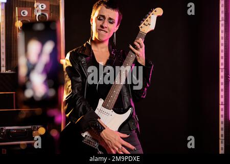 Rebel rockstar tenant une guitare électrique jouant un enregistrement de chanson en métal lourd avec un téléphone moderne pendant une session de musique dans un studio audio. Femme interprète travaillant dans un nouvel album de grunge avant la visite Banque D'Images