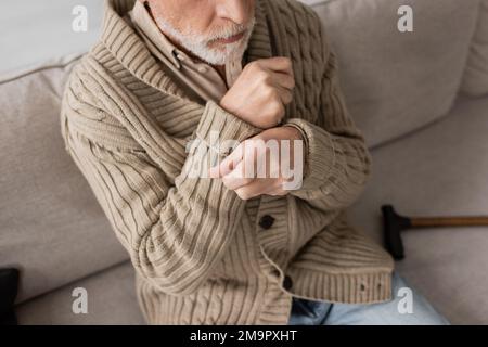 vue courte d'un homme âgé dans un gilet tricoté souffrant d'un parkinsonisme et de tremblements dans les mains lorsqu'il est assis à la maison, image de stock Banque D'Images