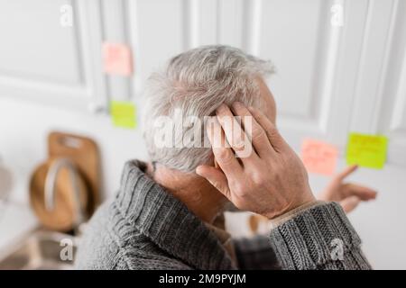 homme âgé à cheveux gris avec le syndrome d'alzheimer touchant la tête et pointant avec un billet collant flou dans la cuisine, image de stock Banque D'Images