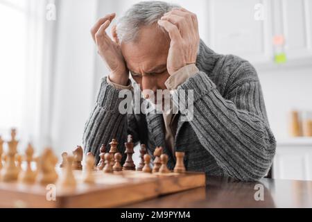 homme stressé souffrant du syndrome d'alzheimer et assis à l'échiquier avec les yeux fermés et les mains près de la tête, image de stock Banque D'Images