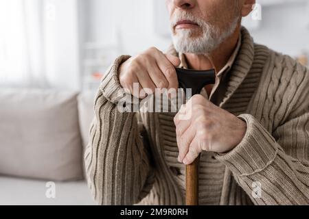 vue partielle de l'homme barbu senior avec le syndrome de parkinson assis avec la canne à pied à la maison, image de stock Banque D'Images