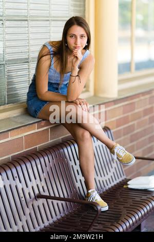 Portrait du corps complet d'une adolescente tendance assise avec des pieds sur un banc à une gare Banque D'Images