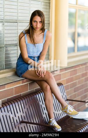 Portrait du corps complet d'une adolescente tendance assise avec des pieds sur un banc à une gare Banque D'Images