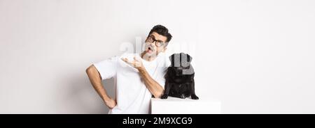 Image d'un type de hipster parlant avec son chien, regardant sceptique et gestante, ayant la conversation avec mignon pug noir, debout sur fond blanc Banque D'Images