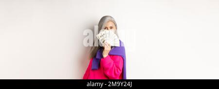 Concept commercial. Une femme coréenne riche et à la mode se cache derrière l'argent, regardant l'appareil photo, debout sur fond blanc Banque D'Images