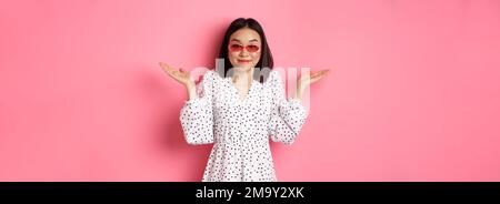 Mignonne asiatique touriste souriant à la caméra, haussant l'indice, ne sais pas, portant des lunettes de soleil à la mode et robe blanche, debout contre fond rose Banque D'Images