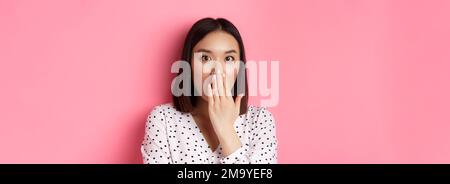 Image d'une fille asiatique choquée, grésillement et bouche de couverture, regarder la caméra avec une incrédulité totale, debout sur fond rose Banque D'Images