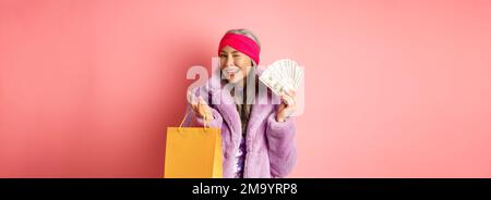 Femme asiatique riche et à la mode gaspillant de l'argent dans les magasins, tenant sac de shopping et dollars, en se faisant un plaisir à l'appareil photo, fond rose Banque D'Images
