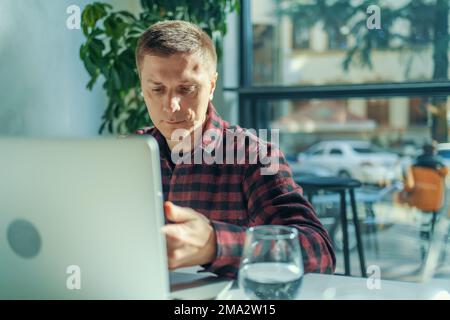 Joyeux indépendant utilisant un ordinateur portable assis à table dans un café par beau temps. Banque D'Images