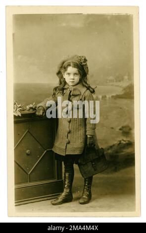 Magnifique original Edwardian Era Studio portrait carte postale de jeune fille édouardienne très mignonne avec de longs cheveux bouclés et portant un manteau tricoté et béret, tenant une mallette en cuir, écolière, circa 1910, Royaume-Uni Banque D'Images