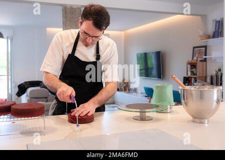 Un boulanger d'homme cuit un gâteau de velours rouge à la maison, coupant le gâteau cuit avec une lyre Banque D'Images