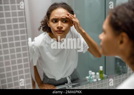 Bouleverser la jeune femme africaine à trouver le bouton sur son visage tout en regardant le miroir dans la salle de bains maison Banque D'Images