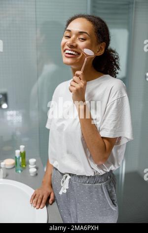 Femme souriante faisant un massage du visage en utilisant un rouleau avec du quartz dans la salle de bains. Concept de cosmétiques naturels Banque D'Images