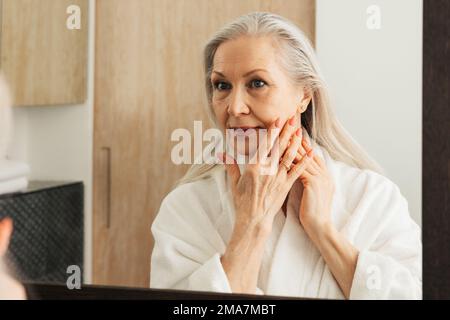 Femme âgée avec de longs cheveux gris touchant sa joue avec les doigts dans la salle de bains Banque D'Images