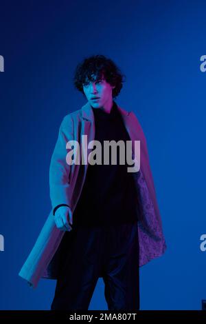 Homme en sèche-cheveux manteau sur fond bleu foncé, lumière néon, style et tendances Banque D'Images