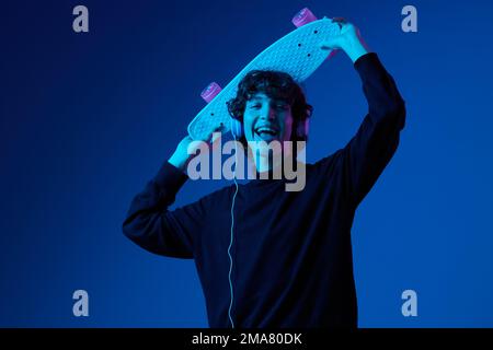 Adolescent avec casque à l'écoute de la musique et danse skateboard à la main sur sa tête, style de vie de hipster, portrait bleu foncé fond, néon Banque D'Images