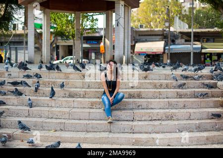 Une belle fille est assise sur les marches du parc entouré de pigeons. Banque D'Images